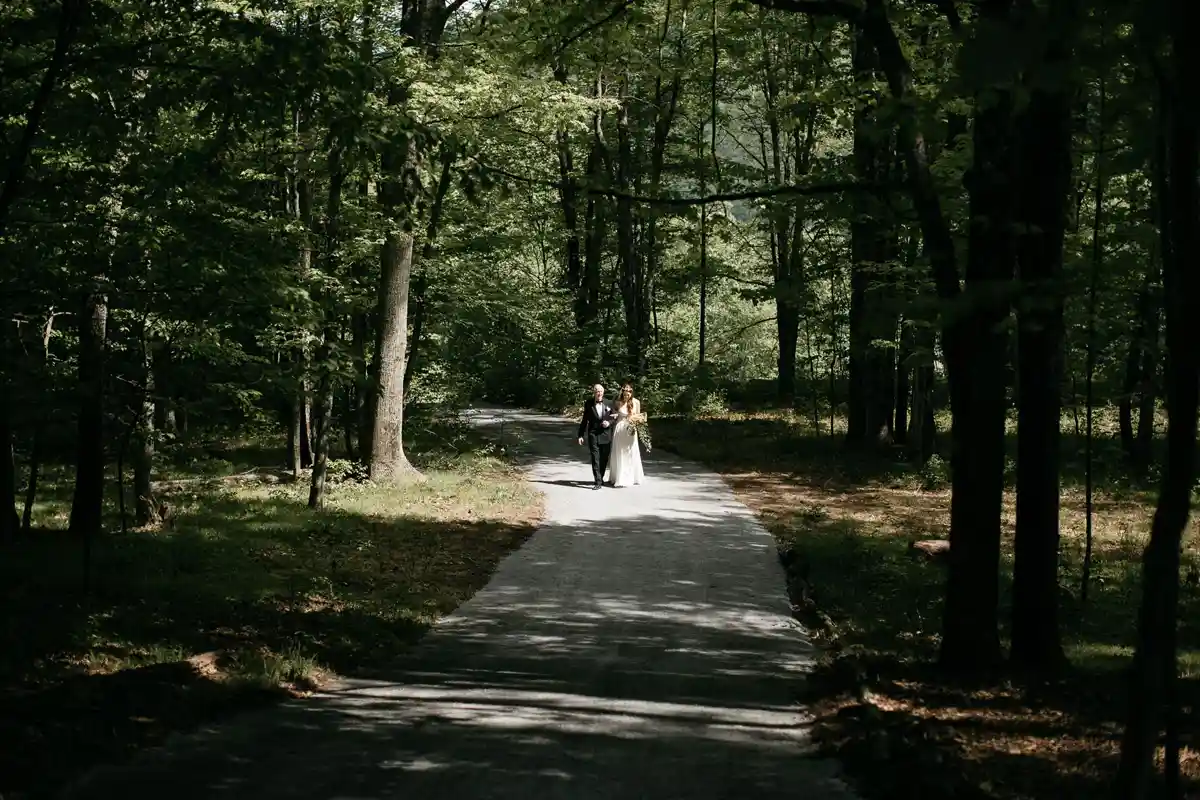 Bride and father walk the aisle on a forest path towards the pine grove at a natural outdoor wedding ceremony location in the Catskills